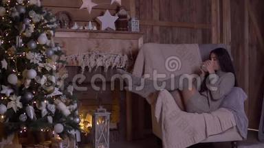 圣诞节，一个女孩坐在圣诞树附近的椅子上喝茶。 高清高清
